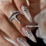 Close-up of a hand with long, almond-shaped nails adorned with intricate silver and clear rhinestone designs, accompanied by silver rhinestone rings.