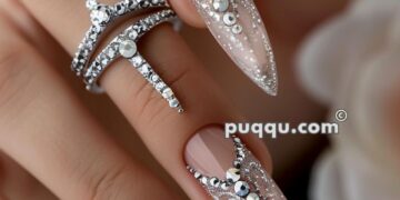 Close-up of a hand with long, almond-shaped nails adorned with intricate silver and clear rhinestone designs, accompanied by silver rhinestone rings.