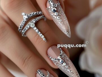 Close-up of a hand with long, almond-shaped nails adorned with intricate silver and clear rhinestone designs, accompanied by silver rhinestone rings.