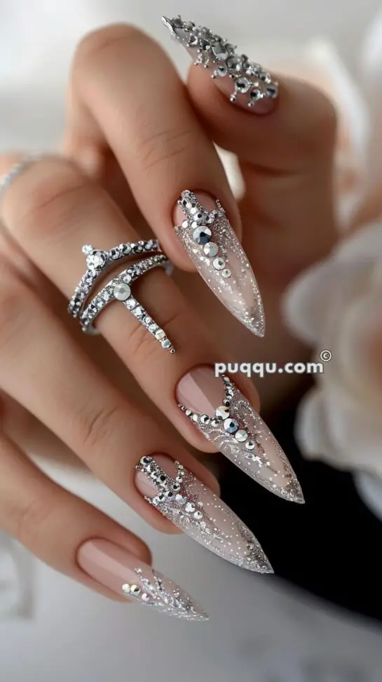 Close-up of a hand with long, almond-shaped nails adorned with intricate silver and clear rhinestone designs, accompanied by silver rhinestone rings.