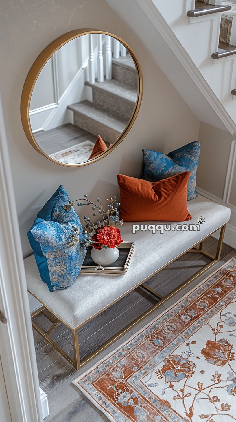 Cozy entryway with a padded bench, adorned with blue and orange decorative pillows and a tray with a flower vase, a round mirror above, and a patterned rug on the floor.