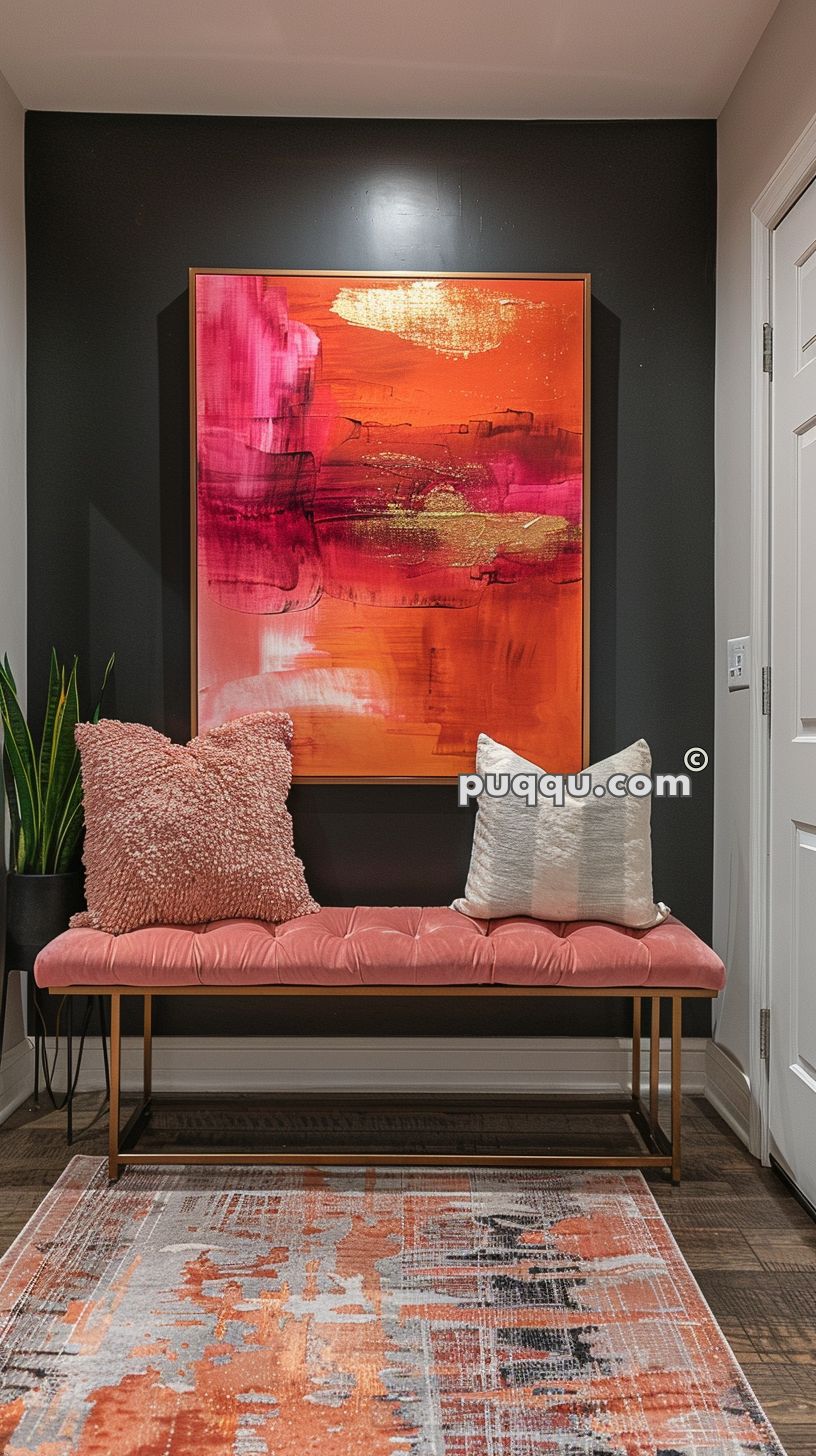 Hallway with dark walls featuring a vibrant abstract painting with red, orange, and gold colors above a cushioned coral bench adorned with two pillows; a textured coral pillow on the left and a white pillow on the right; a potted plant sits to the left of the bench.