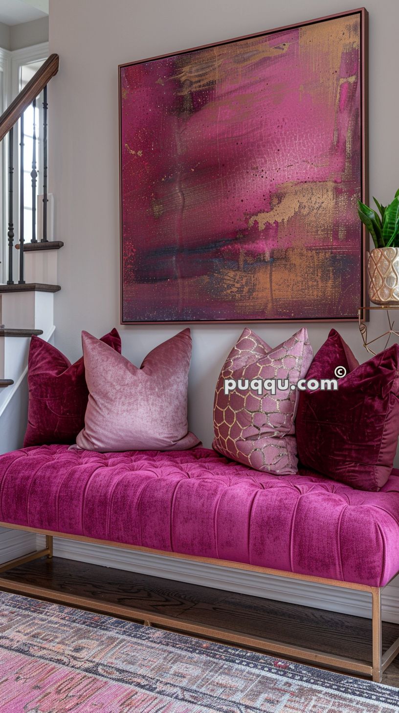 A magenta velvet bench with assorted decorative pillows is placed under a large abstract painting with shades of pink, purple, and gold, adjacent to a staircase.