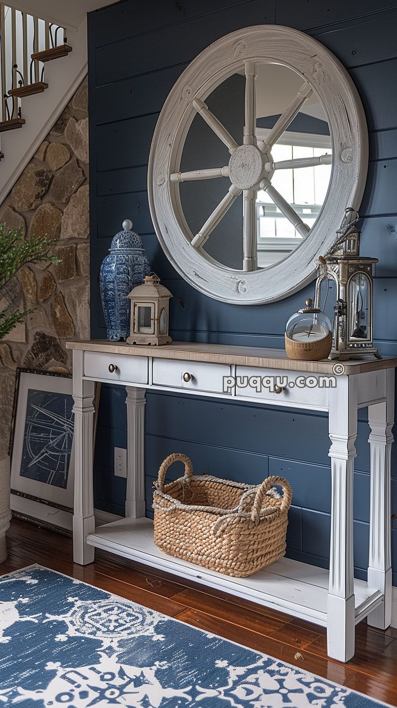 Beach-themed entryway with a white console table, wicker basket, round ship wheel mirror, blue and white ceramic vase, lanterns, framed picture, and blue patterned rug.