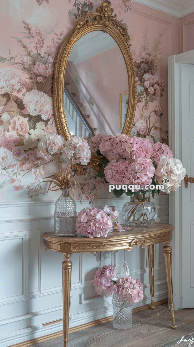 Elegant entryway with a gold ornate mirror above a matching console table, decorated with pink hydrangeas in vases against floral-patterned wallpaper.