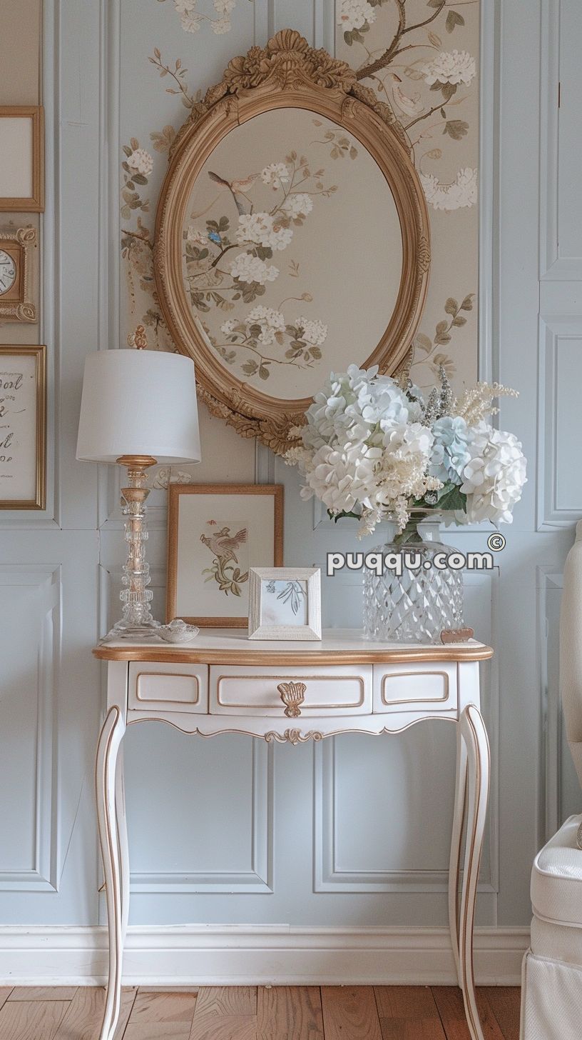 Antique-style console table with a decorative lamp, framed pictures, and a vase of flowers, against a light blue wall with floral wallpaper and an ornate oval mirror.