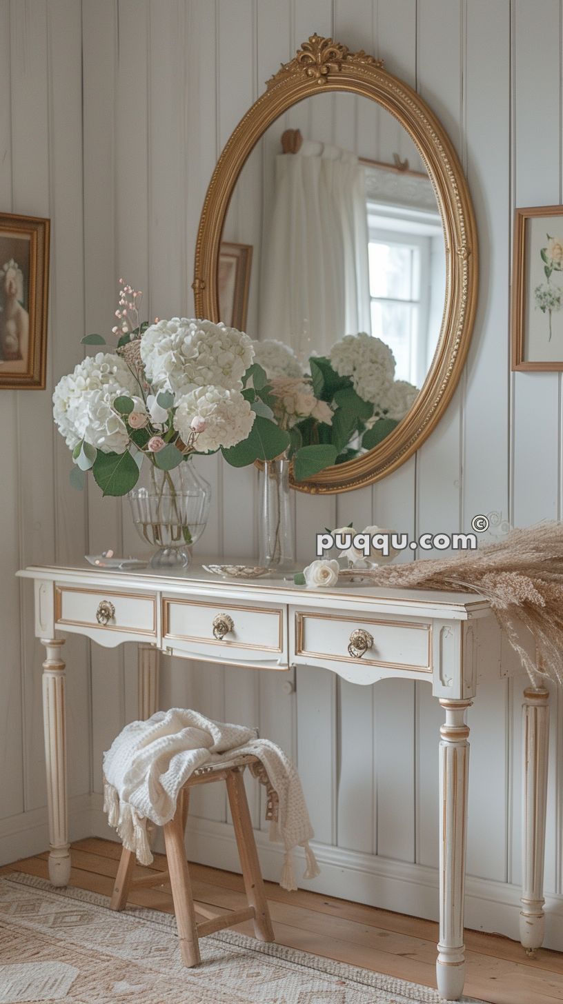 Elegant vanity table with ornate legs and drawer handles, decorated with white hydrangea flowers in glass vases, and a large gilded mirror above. A small wooden stool with a white knitted blanket sits beneath the table.