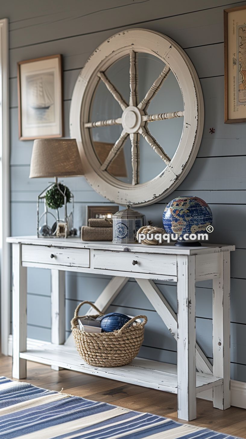 Nautical-themed interior with a distressed white console table, topped with a lamp, globe, and decorative items, set against blue wooden wall paneling. A large ship wheel mirror hangs above, and a striped rug is on the wooden floor.