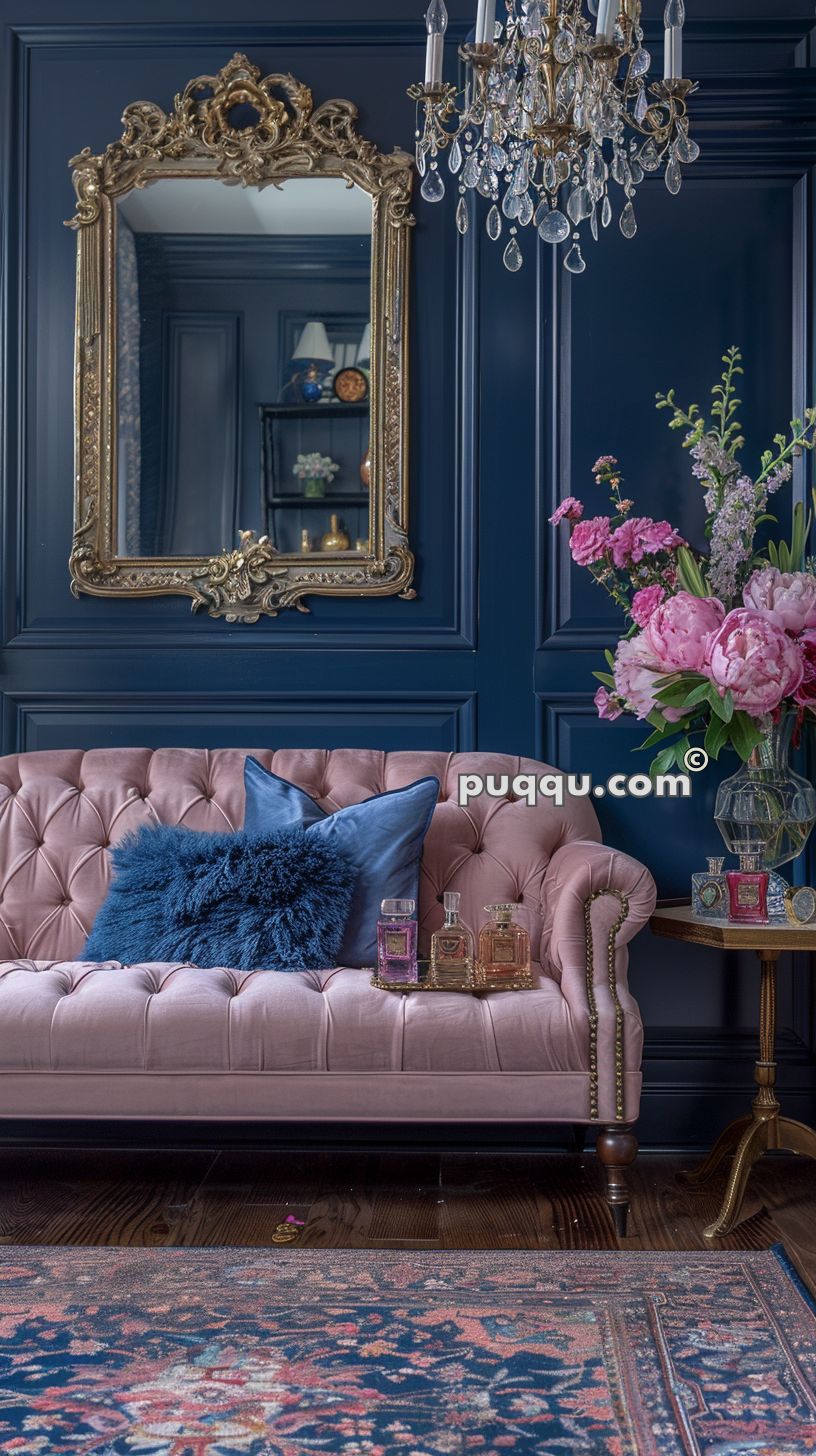 Elegant living room with dark blue walls, ornate gold-framed mirror, pink tufted sofa with blue cushions, crystal chandelier, floral arrangement on a side table, and patterned rug.