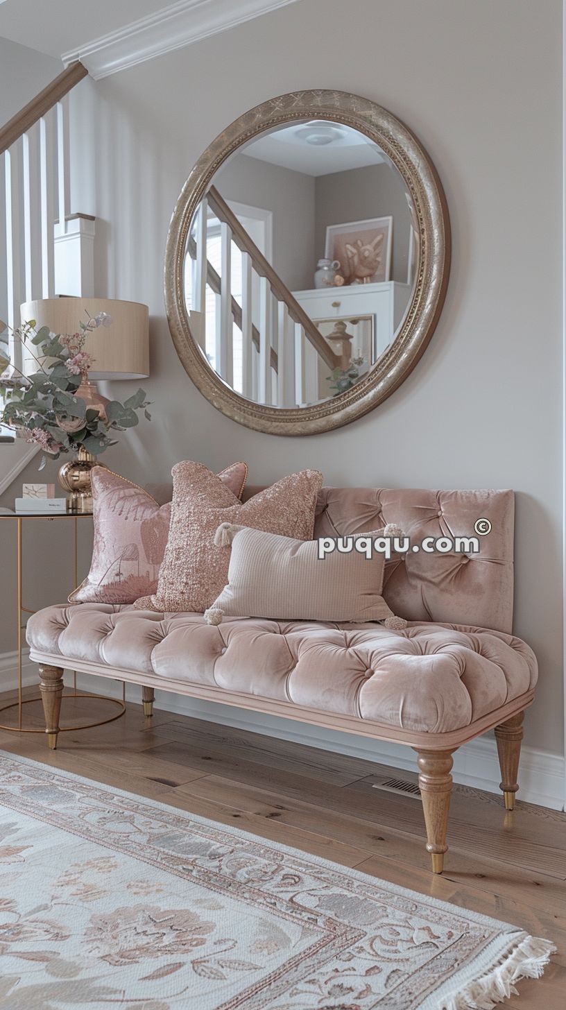 Elegant living space with a pink upholstered bench, decorative pillows, a round wall mirror, and a side table with a lamp and floral arrangement.