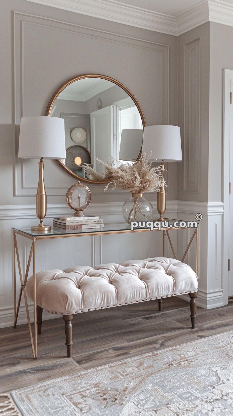 Elegant hallway with a round mirror, gold accents, a glass console table with decorative items, matching gold lamps with white shades, a tufted bench, and light wooden floors.