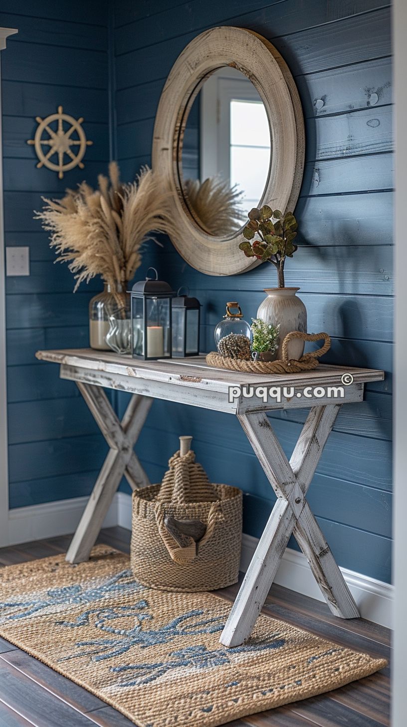 Coastal-themed entryway with a wooden console table decorated with vases, plants, candles, and lanterns, a round mirror hanging above, a jute rug on the floor, and a woven basket below the table, set against blue shiplap walls.