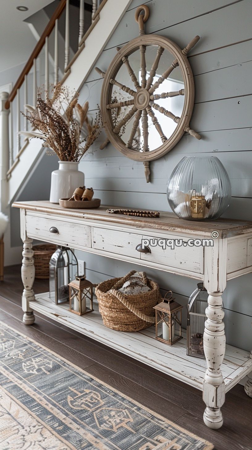 Rustic entryway decor with a whitewashed console table, nautical-themed mirror shaped like a ship's wheel, glass vase, wicker basket, dried flower arrangement, and lanterns.