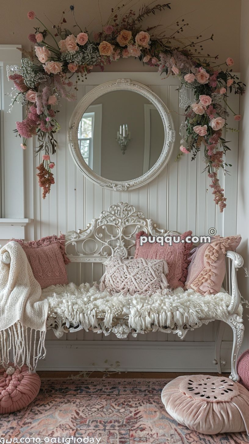 Vintage white bench with decorative pillows and throw, ornate round mirror above, and floral garland in pastel tones hanging over the mirror, set against a beadboard wall.