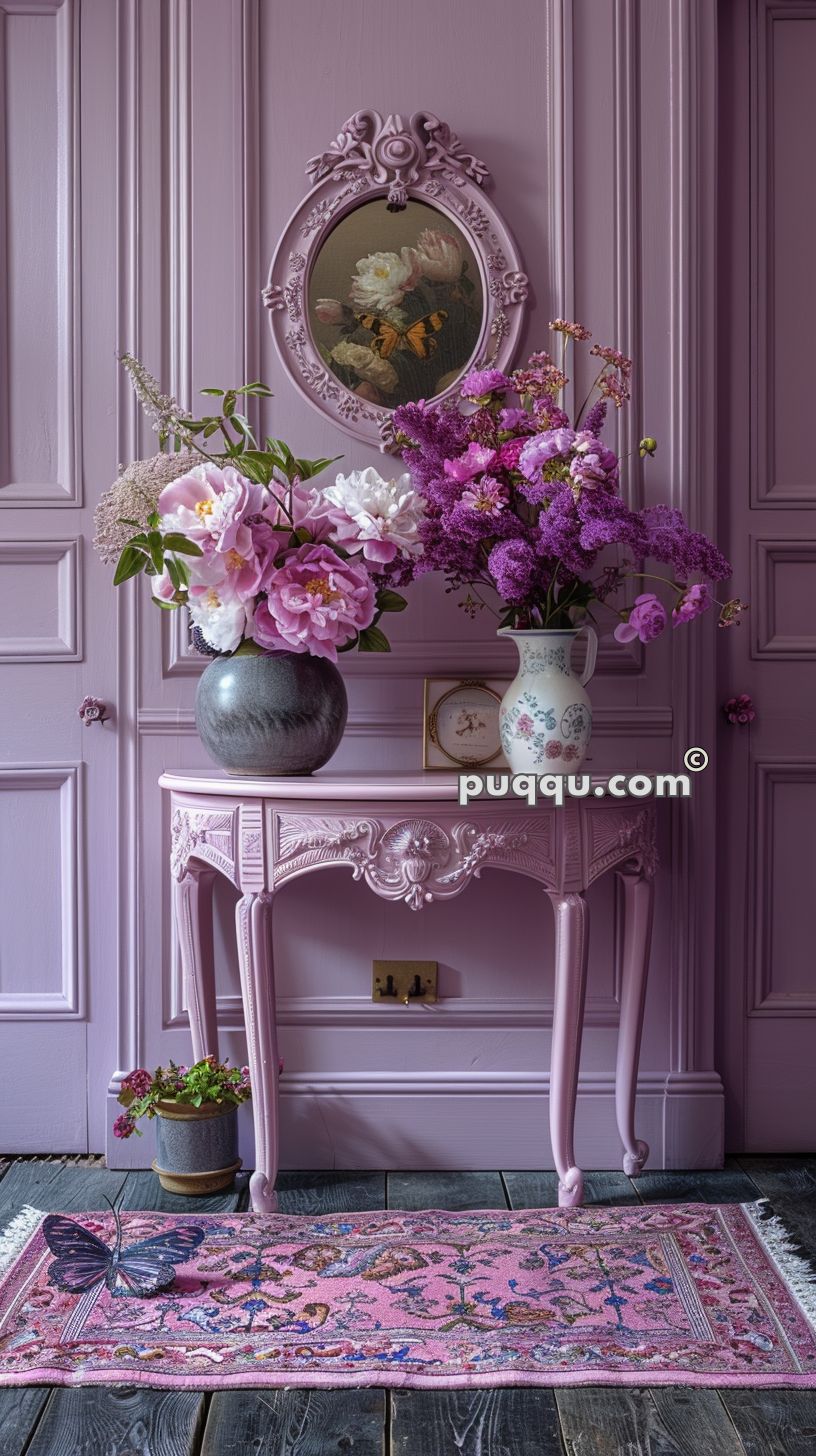 A vintage pink console table with ornate detailing holds two vases with pink and purple flower arrangements, situated against a matching pink paneled wall with an oval floral painting. A colorful rug with a butterfly design lies on the floor in front of the table, and a small potted plant is positioned near the table's legs.