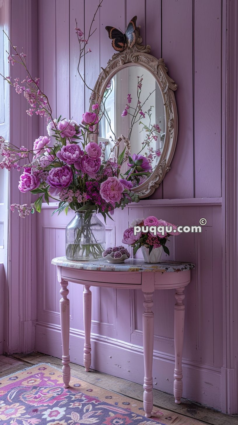 A lavender-colored wooden wall with an ornate oval mirror and butterfly decoration, a table with a glass vase of pink and purple flowers, and a small bouquet in a white vase.