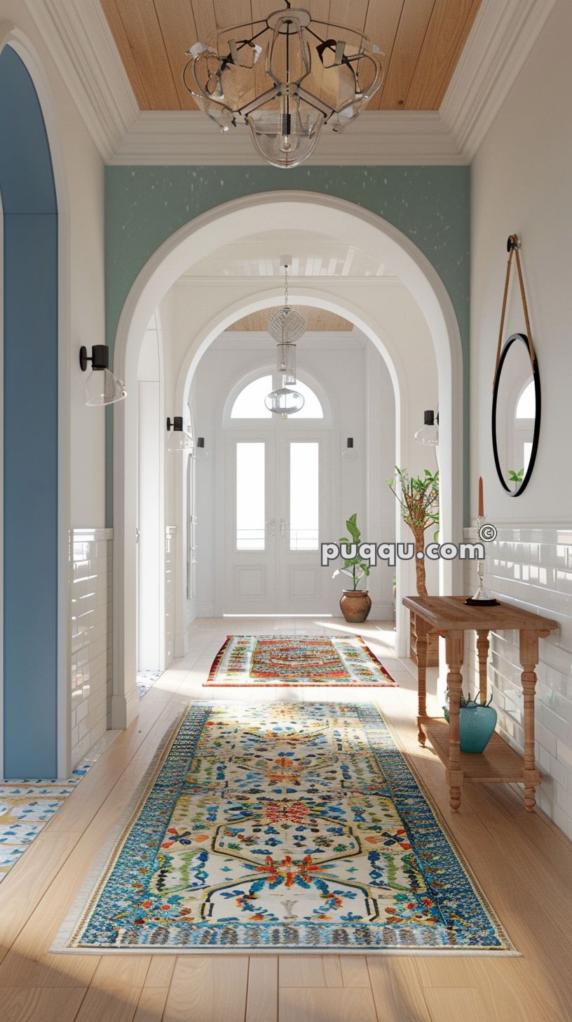 A beautifully decorated hallway with light wood flooring, colorful patterned rugs, white double doors at the end, and an arched ceiling featuring a chandelier. A wooden console table with a round mirror and plant decorates the right side.