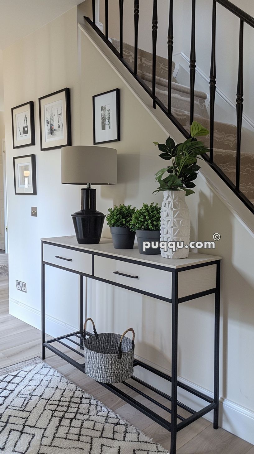 Modern entryway with a console table featuring a black lamp, small plants, and a decorative white vase. Framed artwork adorns the wall above, and a basket rests on the lower shelf of the table.