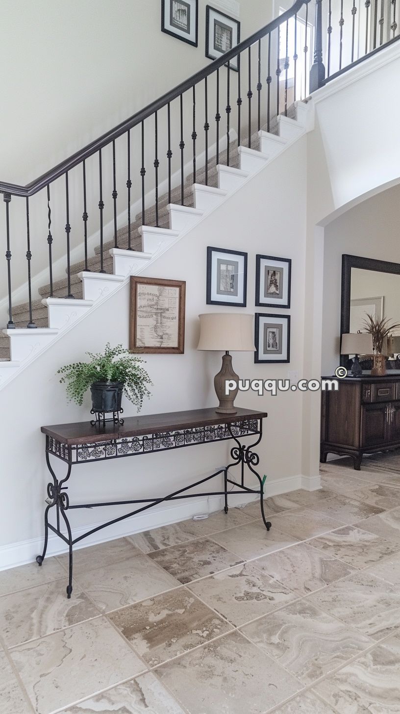 Staircase with dark metal railings, a tile floor, and a decorative console table with a lamp and potted plant underneath framed wall art.