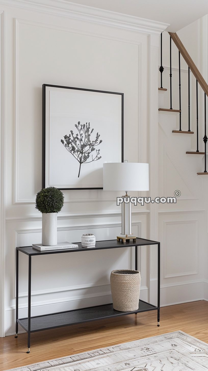 Minimalist entryway with a black console table, a small potted plant, a decorative jar, and a white table lamp. A framed black and white botanical print hangs above the table, with a staircase visible on the right.