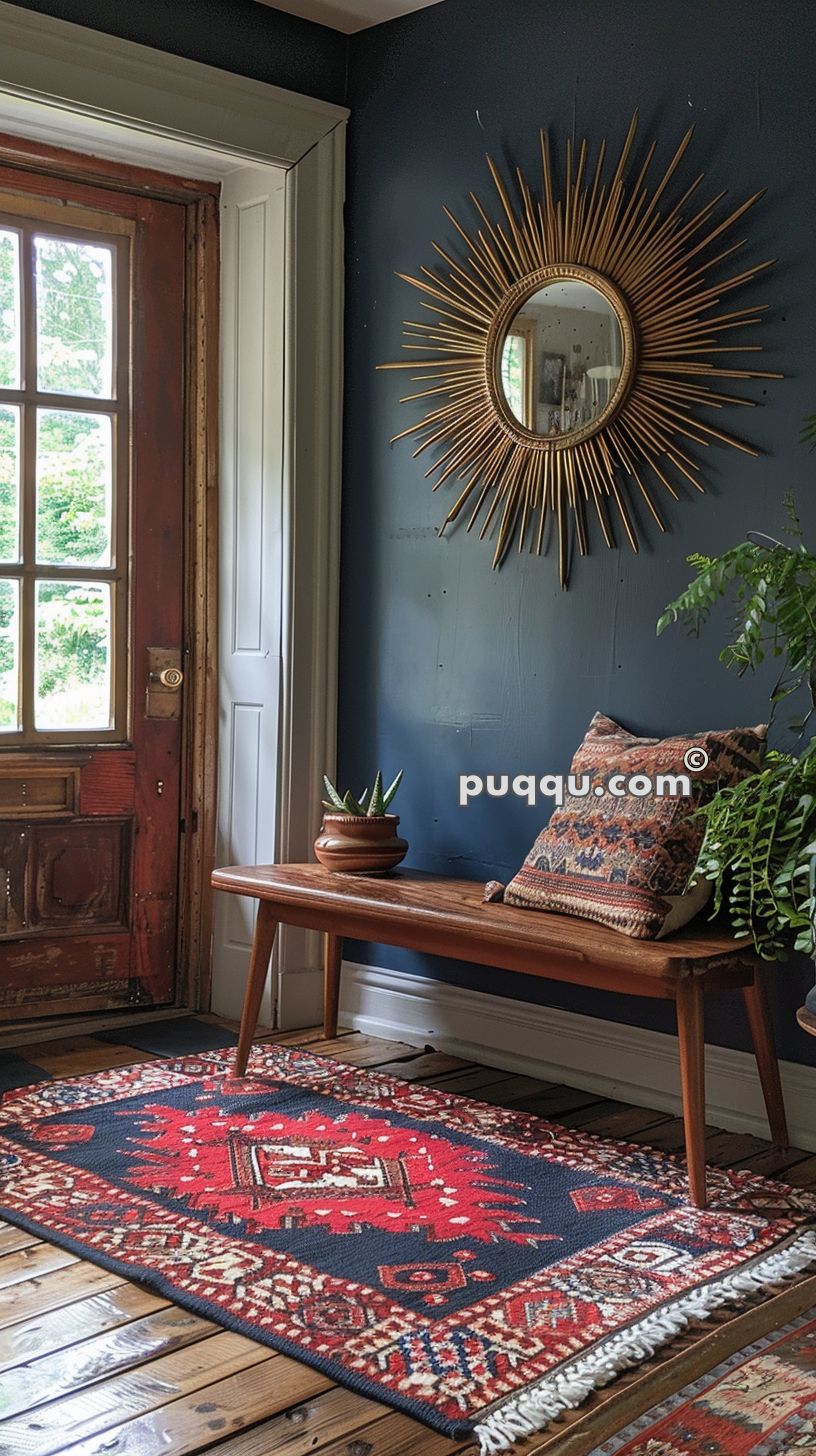 Entryway with wooden door, large window, wooden bench, decorative pillow and plant, navy blue wall with golden sunburst mirror, and a red and navy patterned rug on wooden floor.