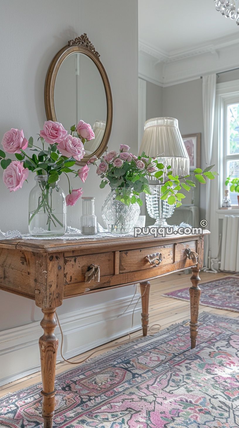 Antique wooden table with pink roses in glass vases, an oval wall mirror, a table lamp, and decorative items in a bright room with patterned rugs.