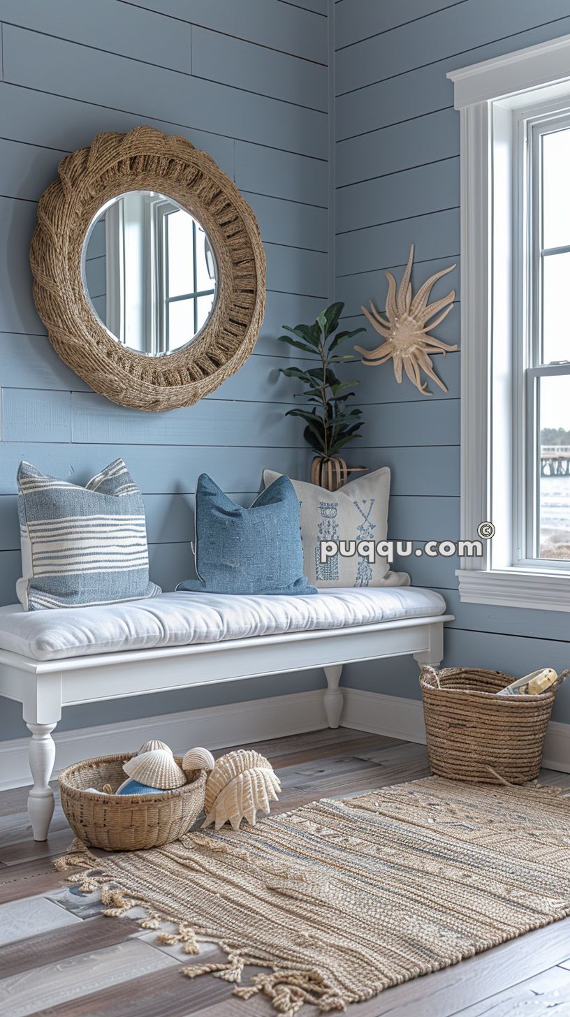 Cozy beach-themed corner with a white bench, blue and white throw pillows, a woven mirror, a seashell wall decor, and a woven rug. Baskets and seashells are placed nearby.