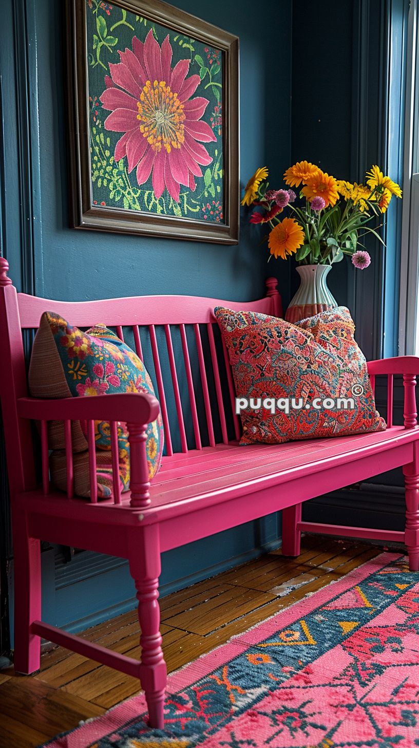 Bright pink wooden bench with colorful floral cushions placed against a dark blue wall, featuring a large framed painting of a pink flower. A bouquet of vibrant flowers in a vase sits on a side table near the bench, next to a window.