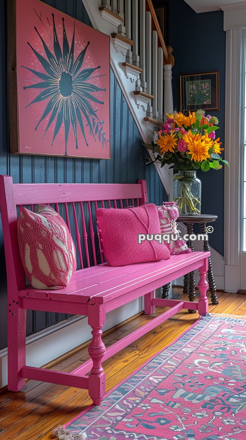Pink wooden bench with decorative pillows under a large floral painting in a bright, colorfully decorated entryway with wooden floors and a patterned rug.
