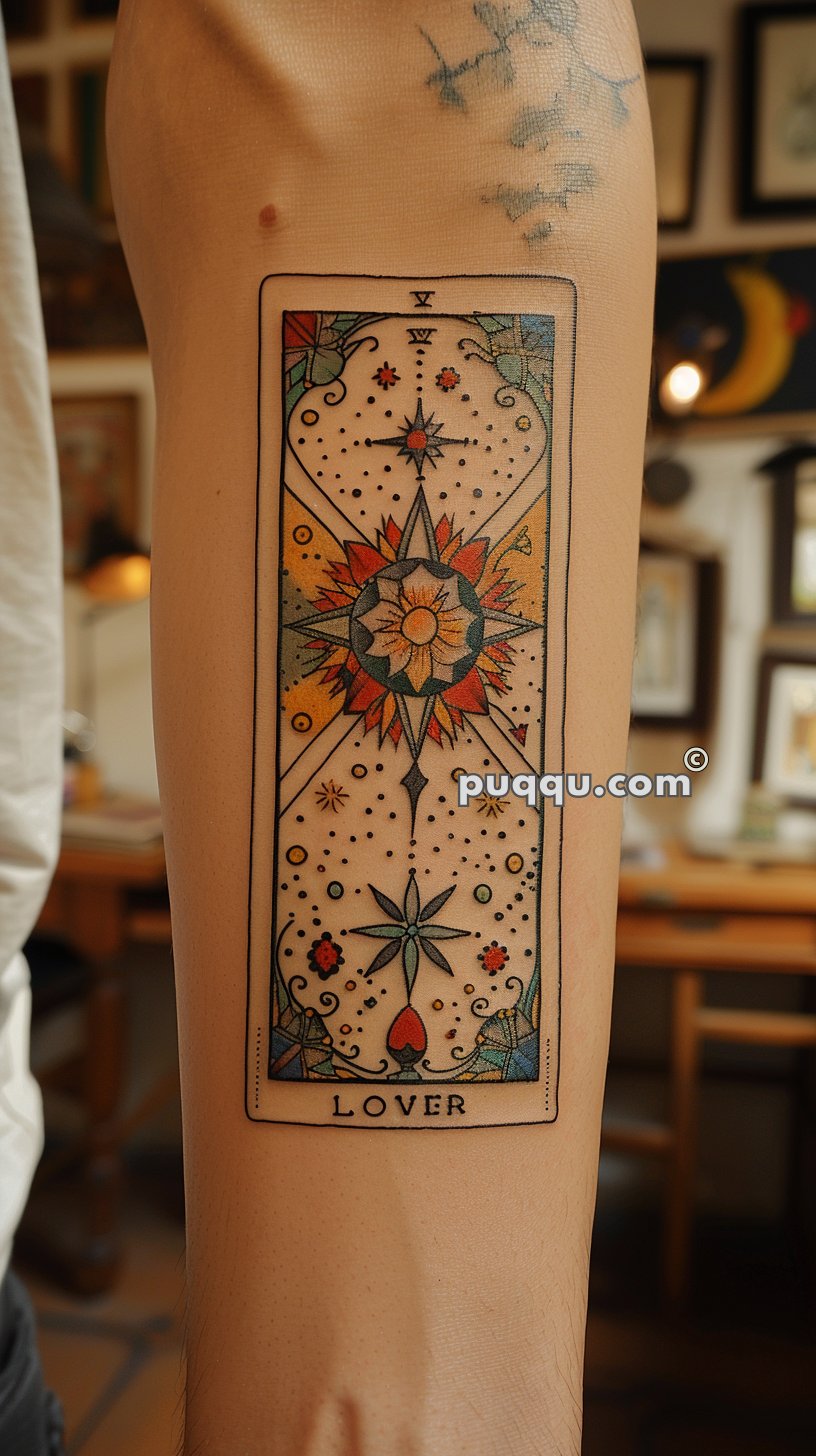 A colorful tattoo on the inner forearm depicting a tarot card design with intricate geometric and floral patterns, along with the word "LOVER" at the bottom.