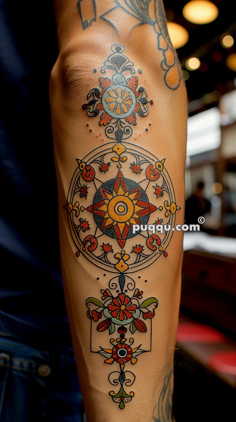 Intricate mandala tattoo design on an arm, featuring a central starburst pattern surrounded by floral and ornamental details in red, yellow, and green.