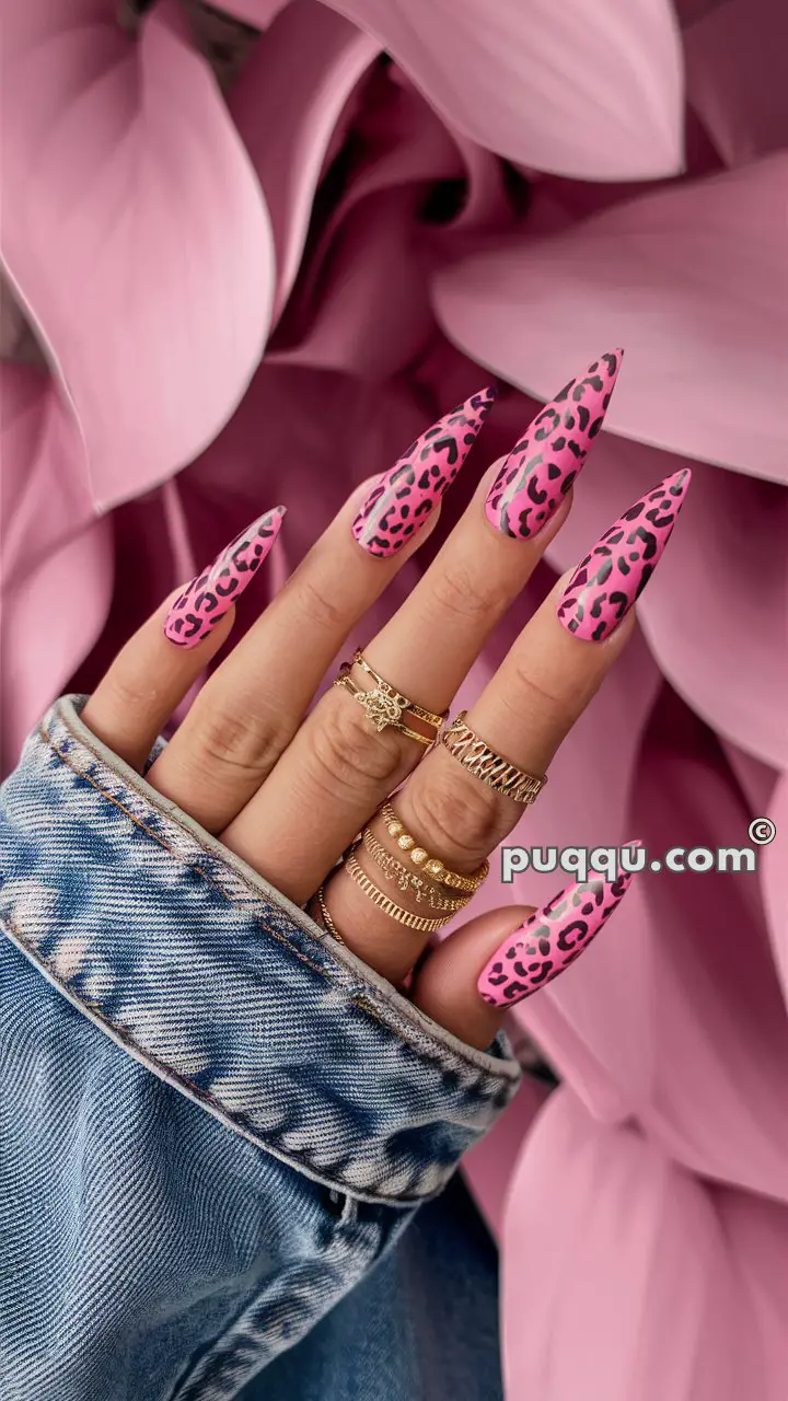 A hand with long pink and black leopard print nails, adorned with multiple gold rings, against a background of large pink leaves.