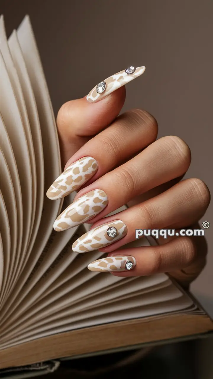 Close-up of a hand with long, pointed nails featuring a beige and white giraffe print design, adorned with jeweled accents, holding an open book.