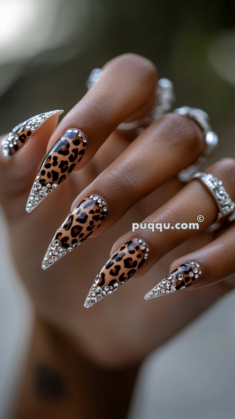 Close-up of a hand with long, pointed nails featuring leopard print and rhinestone accents, wearing multiple silver rings.