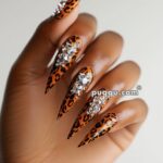 Hand with long, pointed nails featuring leopard print and adorned with large rhinestones.