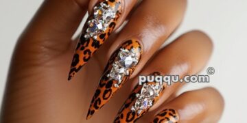 Hand with long, pointed nails featuring leopard print and adorned with large rhinestones.