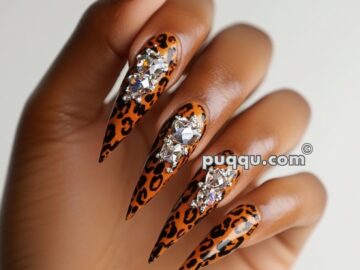 Hand with long, pointed nails featuring leopard print and adorned with large rhinestones.