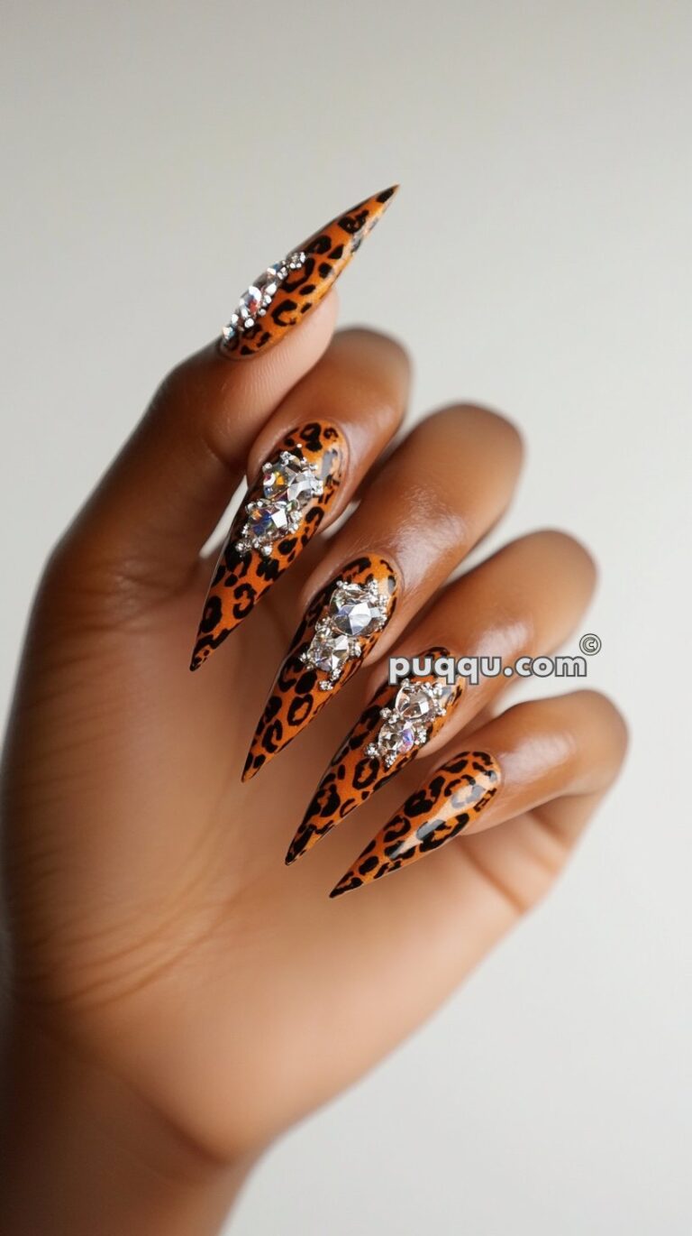 Hand with long, pointed nails featuring leopard print and adorned with large rhinestones.