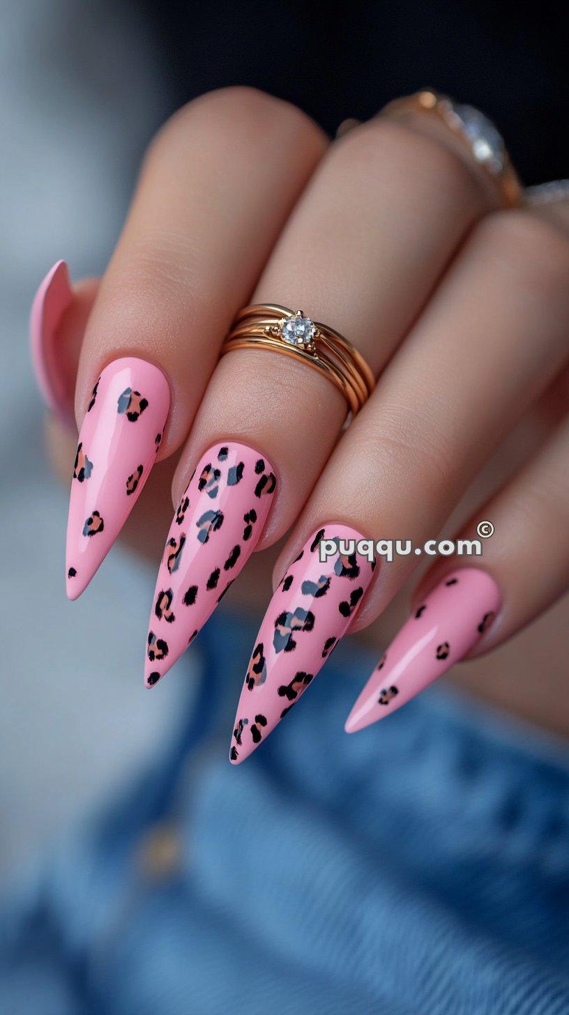 Hand with long, pointed nails painted in pink with black leopard print accents, wearing gold rings with a diamond.