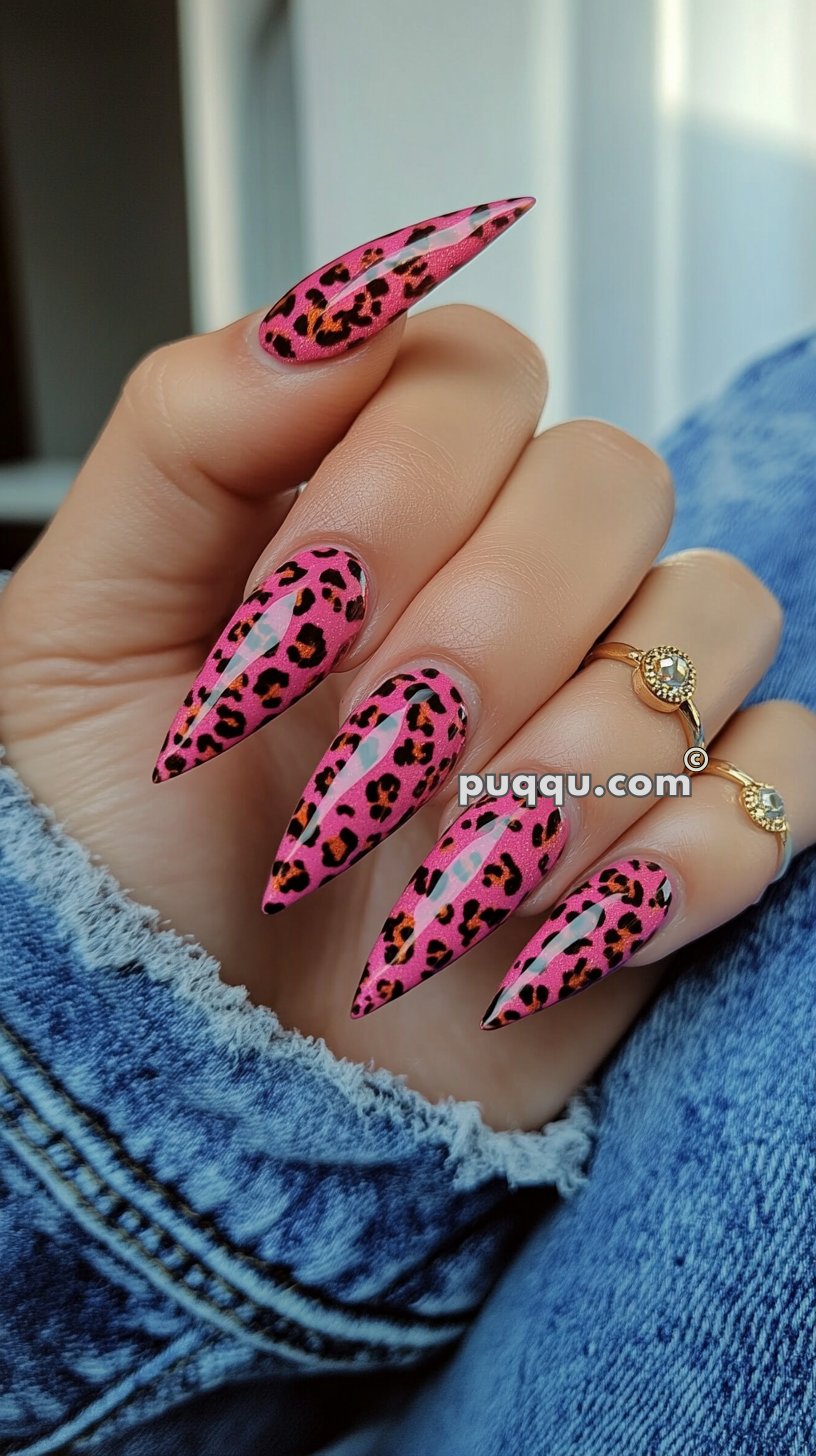 A hand with long, pointed nails featuring a pink leopard print design, with gold rings on the fingers and wearing a denim jacket.