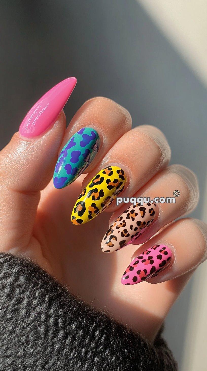 A hand with almond-shaped nails, each painted with a different bold design: plain pink, blue with purple spots, yellow with black and orange spots, nude with brown and black spots, and pink with black spots.