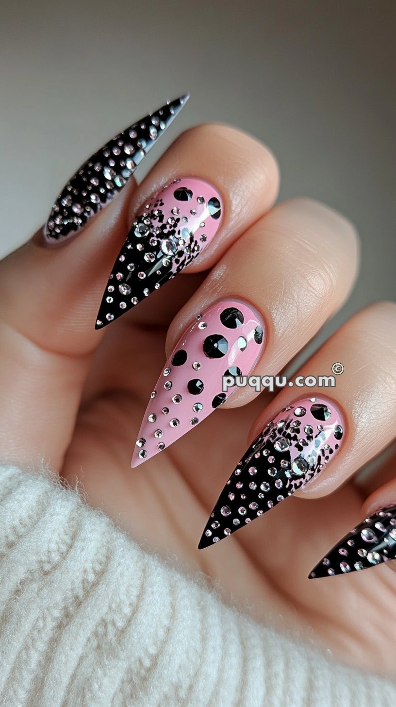 Close-up of hand with long, pointed nails featuring a pink and black design adorned with rhinestones and polka dots.