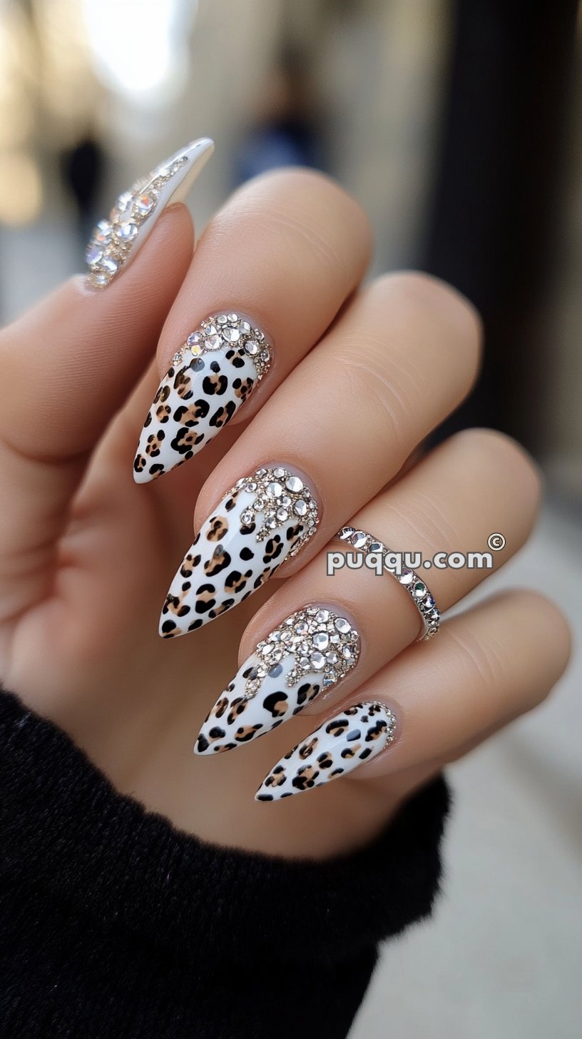 Close-up of a hand displaying stiletto-shaped nails with a white base, leopard print design, and rhinestone embellishments near the cuticles.