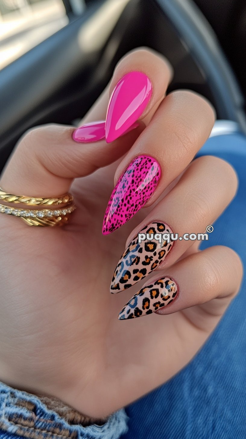 Hand with long stiletto-shaped nails, featuring a mix of bright pink, pink with black specks, and leopard print designs. The fingers have gold and diamond rings.