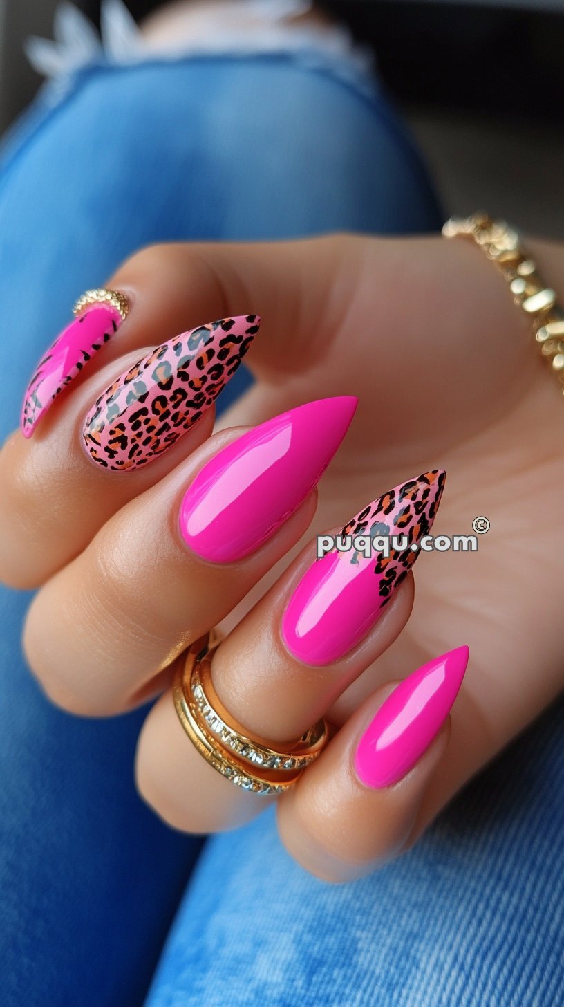 Bright pink stiletto nails with leopard print accents, gold rings on the fingers, and blue denim background.