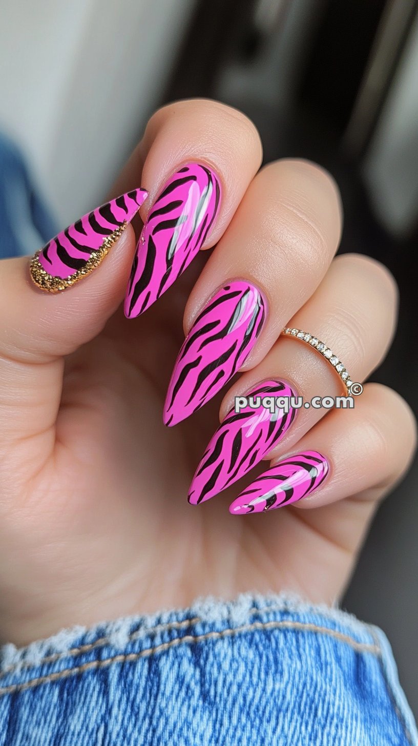 Pink stiletto nails with black zebra stripes and gold glitter accents on one nail, paired with a rhinestone ring.