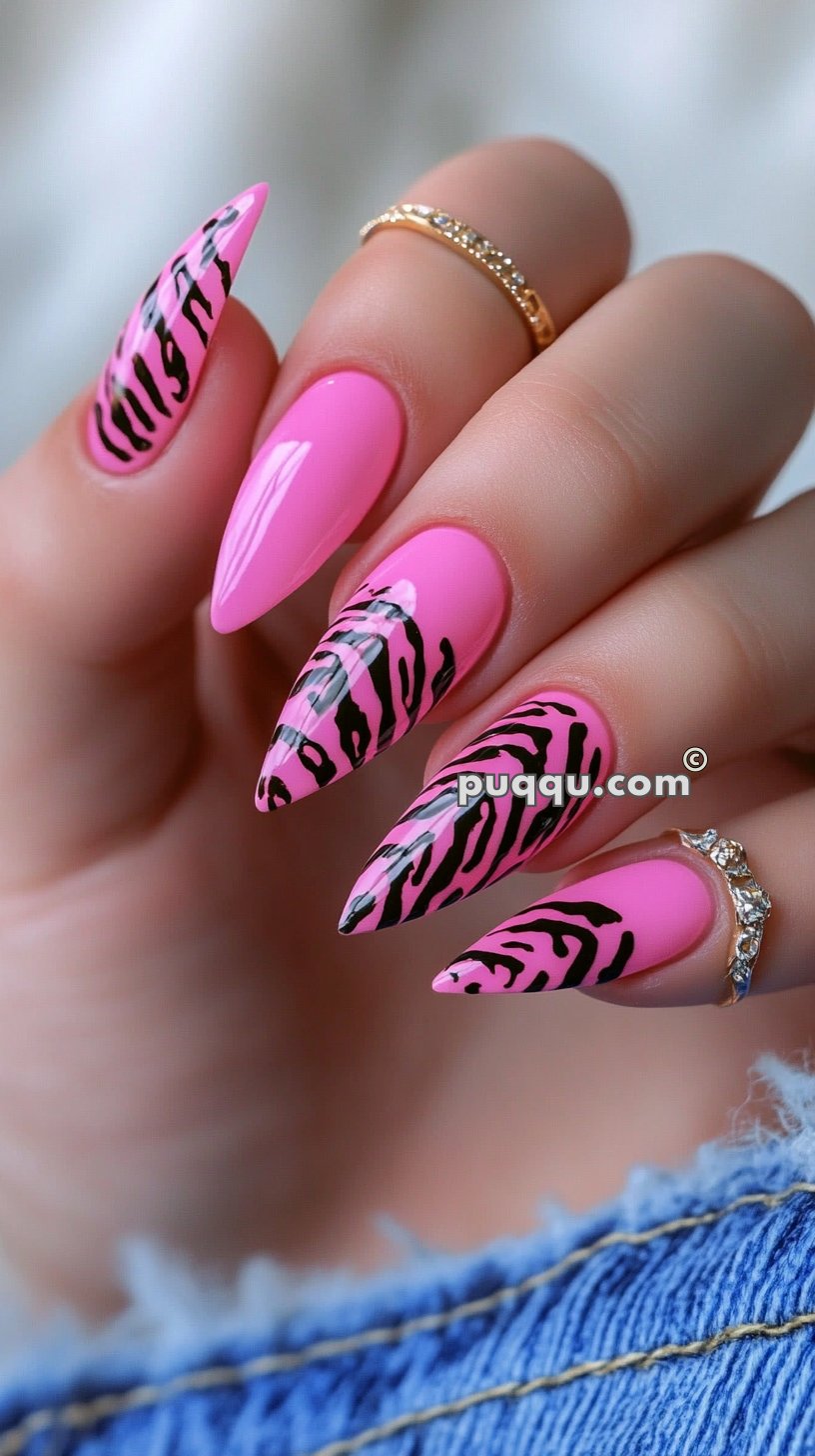 Hand with long, pointed nails painted in bright pink and black zebra print design, wearing gold rings with small diamonds.