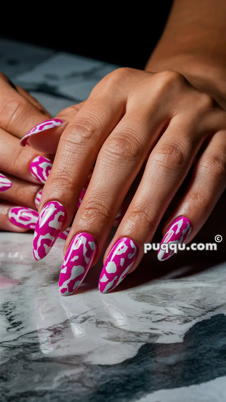 Hands with long, almond-shaped nails painted in a pink and white cow spot pattern on a marble surface.