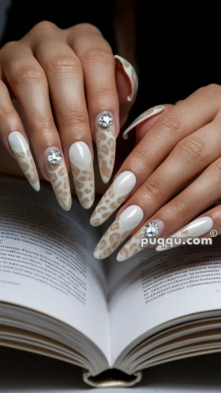 Hands with long nails painted in a beige and white leopard print design, adorned with rhinestones, holding an open book.