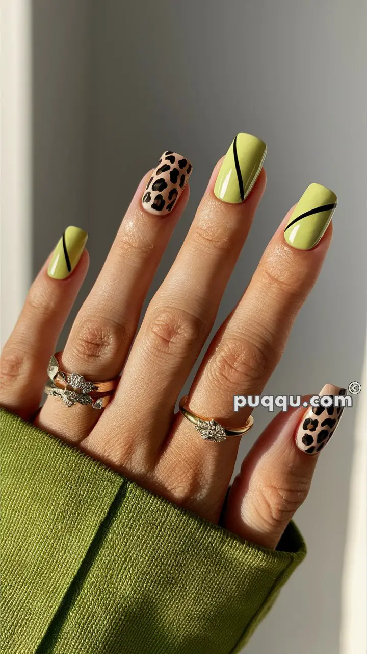 A hand with manicured nails featuring lime green polish with black diagonal stripes and leopard print designs, wearing three rings with floral and solitaire designs.